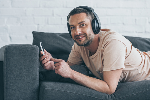 cheerful man in headphones  while lying on sofa and using smartphone with blank screen