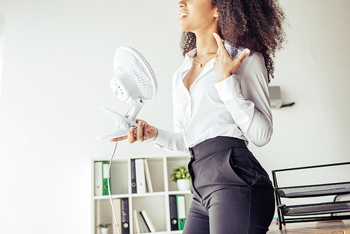 partial view of african american businesswoman holding desk fan while suffering from heat in office