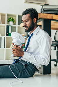 pensive african american businessman holding electric fan while sitting on floor and suffering from summer heat