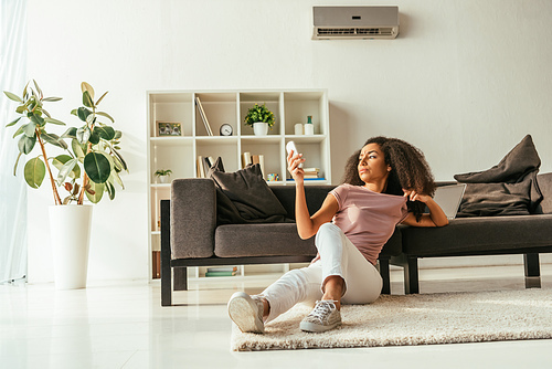 attractive african american woman sitting on floor and using air conditioner remote controller while suffering from summer heat at home