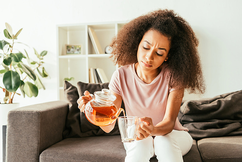 young african american woman pouring tea into cup while sitting on sofa and suffering from heat at home