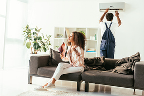 pretty african american woman sitting on couch and waving with magazine, while african american repairman fixing air conditioner