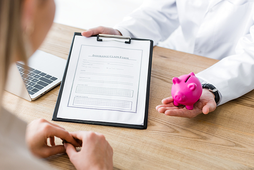 cropped view of doctor holding insurance claim form and piggy bank in hand