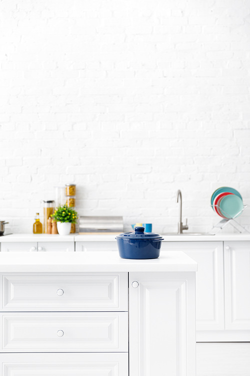 selective focus of blue pot and minimalistic modern white kitchen interior with kitchenware and food near brick wall on background