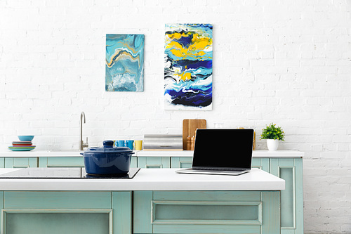 selective focus of laptop and pot on induction cooktop with kitchenware and abstract paintings on background
