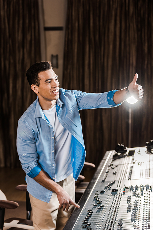 cheerful mixed race sound producer showing thumb up while working at mixing console