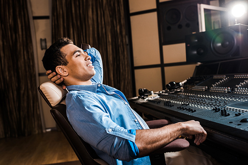 smiling mixed race sound producer relaxing in office chair in recording studio
