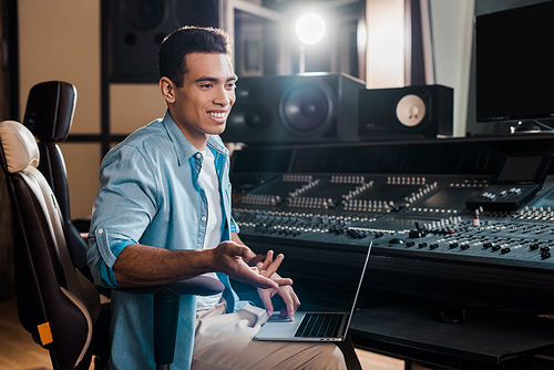 smiling mixed race sound producer sitting by mixing console and using laptop