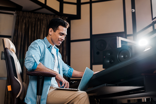 smiling mixed race sound producer sitting in recording studio and using laptop