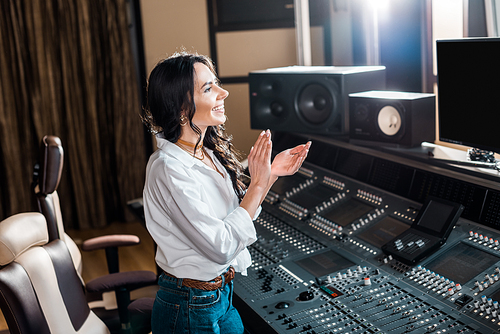 attractive, smiling sound producer applauding in recording studio near mixing console