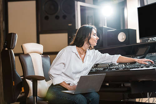 attractive, smiling sound producer using laptop and working at mixing console in recording studio