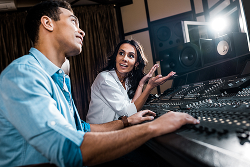 selective focus of pretty sound producer gesturing near mixed raced colleague working at mixing console