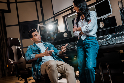 two young multicultural sound producers talking near mixing console in recording studio