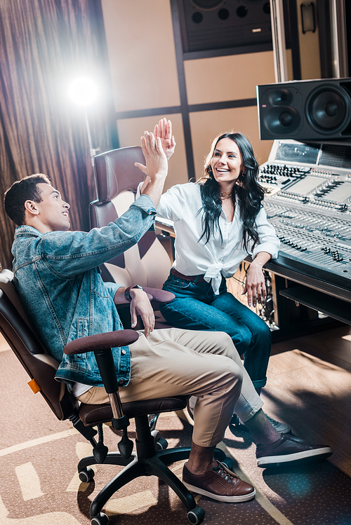 two cheerful multicultural sound producers giving five in recording studio while sitting near mixing console