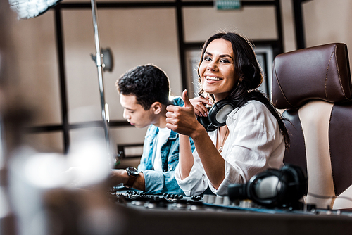 selective focus of smiling sound produce showing thumb up while working at mixing console near mixed race friend