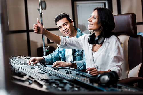 selective focus of cheerful sound produce showing thumb up while working at mixing console near smiling mixed race friend