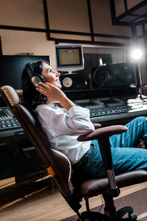 pretty, smiling mixed raced sound producer listening music in headphones near mixing console