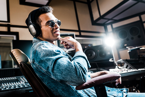 smiling mixed race sound producer in headphones and sunglasses sitting in recording studio