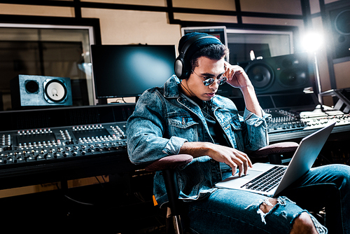 concentrated mixed race sound producer in headphones using laptop in recording studio
