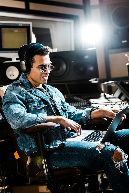 smiling mixed race sound producer in headphones using laptop in recording studio