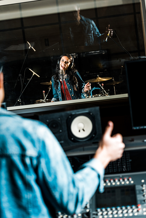 selective focus of mixed race sound producer showing thumb up to beautiful singer in recording studio