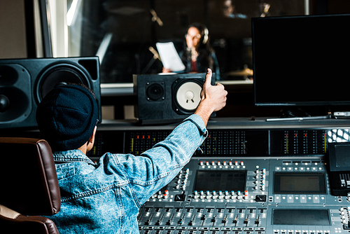 selective focus of mixed race sound producer showing thumb up to woman singing in recording studio