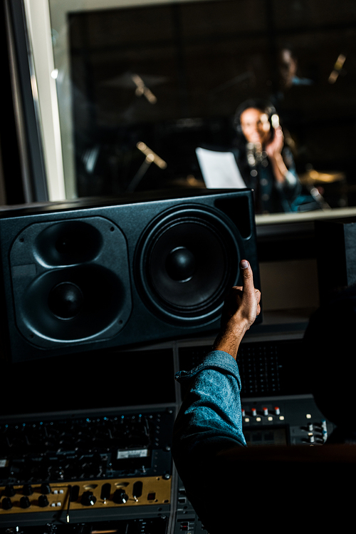 selective focus of mixed race sound producer showing thumb up to woman singing in recording studio