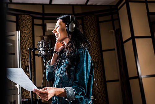 selective focus of pretty woman holding paper with text while singing in recording studio