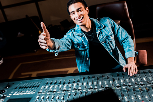 smiling mixed race sound producer showing thumb up while standing near mixing console in recording studio