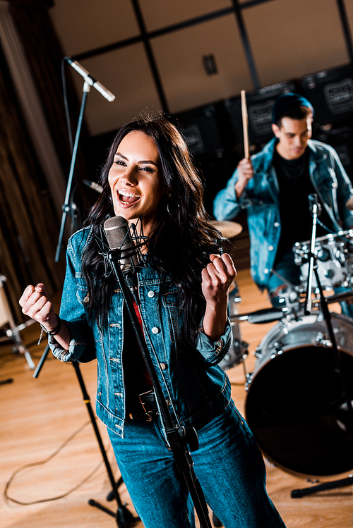 selective focus of beautiful emotional woman singing while mixed race musician playing drums in recording studio