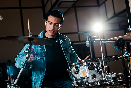 handsome mixed race musician playing drums in dark recording studio