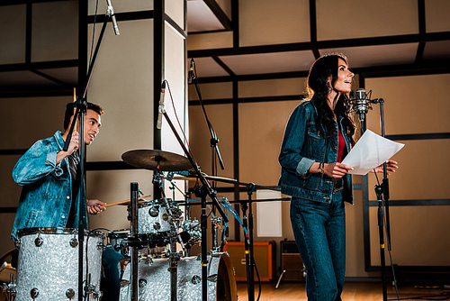 handsome mixed race musician paying drums and attractive woman singing in recording studio
