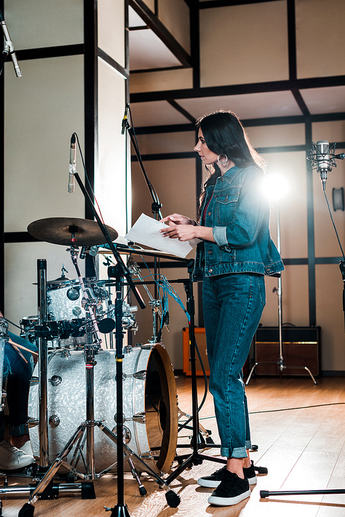 attractive singer standing near microphone and holding paper with text in recording studio