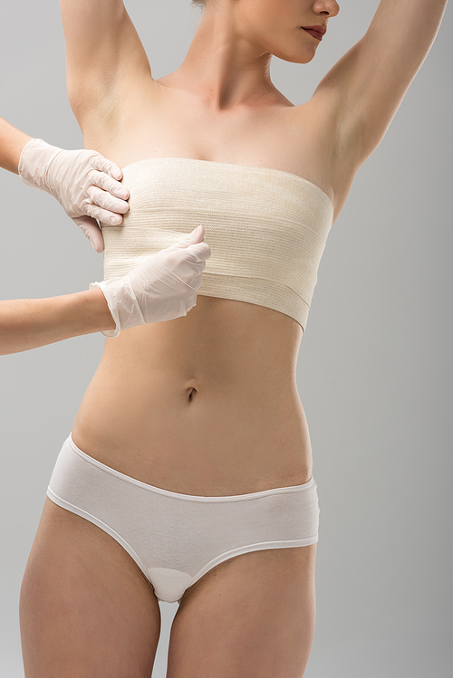 partial view of plastic surgeon in latex gloves and patient in breast bandage isolated on grey