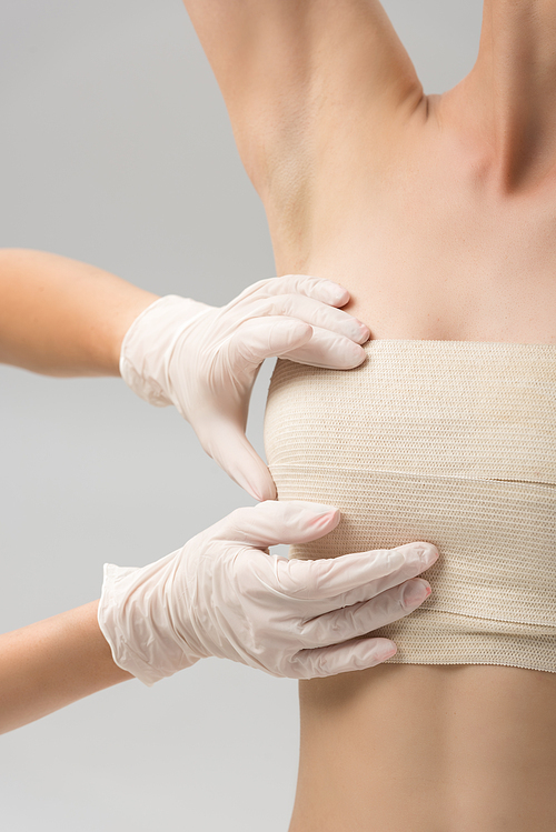 partial view of plastic surgeon in latex gloves and patient in breast bandage isolated on grey