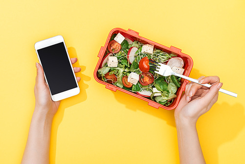 cropped view of woman with fork and lunch box with salad using smartphone