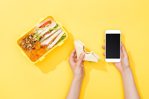 cropped view of woman holding sandwich and smartphone near lunch box with food