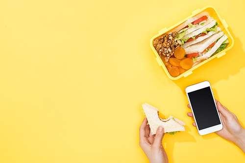 cropped view of woman holding sandwich and smartphone near lunch box with food