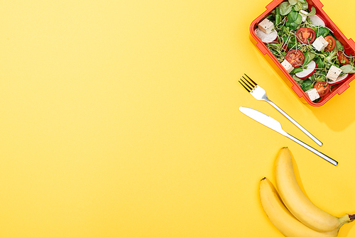 top view of bananas, fork and knife near lunch box with salad