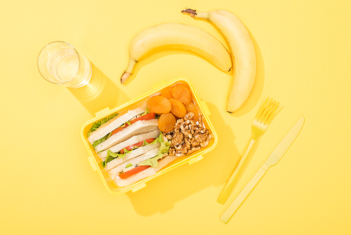top view of lunch box with food near glass of water, bananas and plastic utensils