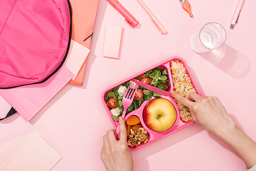 cropped view of woman hands with plastic utensils over lunch box with food near backpack and stationery