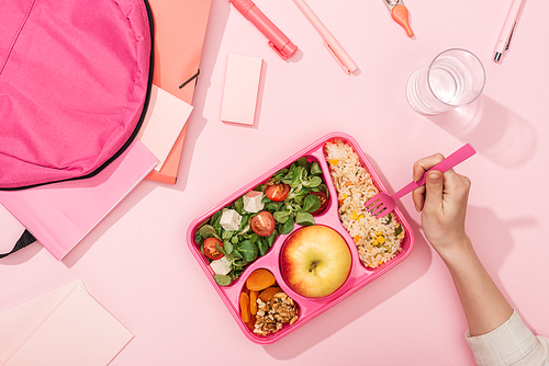 cropped view of woman hands with plastic utensils over lunch box with food near backpack and stationery