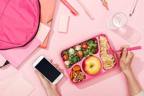 cropped view of woman with smartphone near lunch box, backpack and stationery
