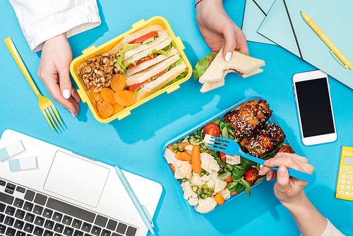 cropped view of two women holding forks over lunch boxes with food near laptop