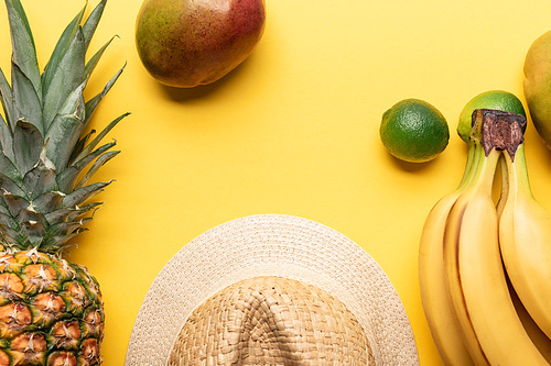 top view of straw hat near pineapple, bananas, limes and mango on yellow background