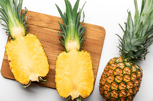 top view of cut ripe yellow pineapple on wooden chopping board near whole fruit on white background