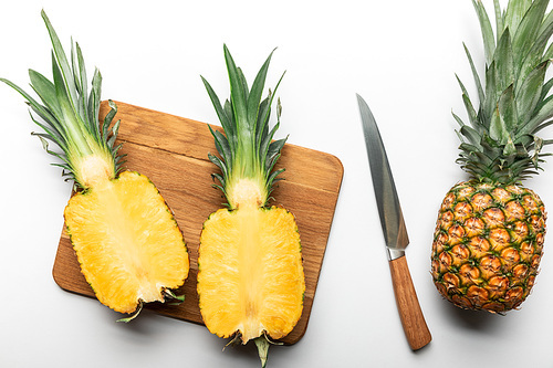 top view of cut ripe yellow pineapple on wooden chopping board near whole fruit and knife on white background