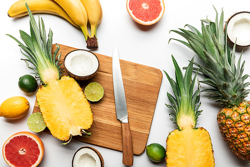 top view of cut and whole tropical fruits on wooden chopping board near knife on white background