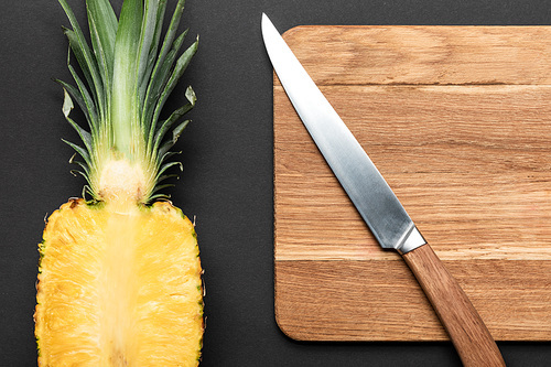 top view of ripe pineapple half on wooden chopping board with knife on black background