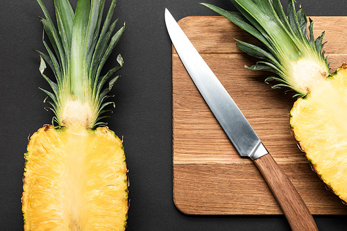 top view of cut ripe pineapple on wooden chopping board with knife on black background